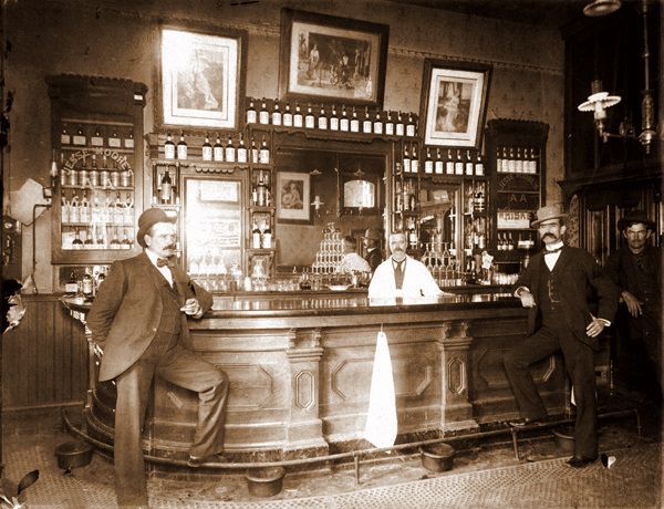 Seepiansävyisessä kuvassa on vanha amerikkalainen baari, jossa tiskin edessä seisoo viiksekkäitä knallipäiviä miehiä poseeraamassa.