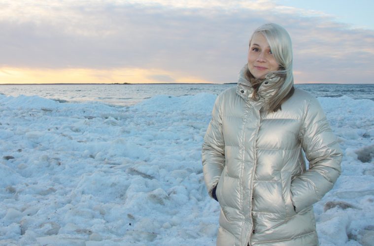 Vaaleahiuksinen Jenny Simojoki on pukeutunut kullanväriseen untuvatakkiin ja katsoo kameraan kädet taskussa. Takana on talvista maisemaa, meri ja auringonlaskun värejä.